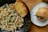 A close-up of a meal on a wooden table. On a black speckled plate, there is creamy pasta with fusilli and mixed vegetables garnished with parsley, accompanied by a slice of toasted garlic bread. To the side, a white plate holds a sesame seed bun with a green chili pepper placed on top of it.