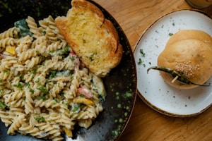A close-up of a meal on a wooden table. On a black speckled plate, there is creamy pasta with fusilli and mixed vegetables garnished with parsley, accompanied by a slice of toasted garlic bread. To the side, a white plate holds a sesame seed bun with a green chili pepper placed on top of it.