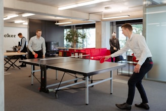 Two individuals are engaged in a game of table tennis in an office setting. The room is well-lit with modern furnishings including a red couch and potted plants. In the background, there are other people working at desks and someone wearing a VR headset.