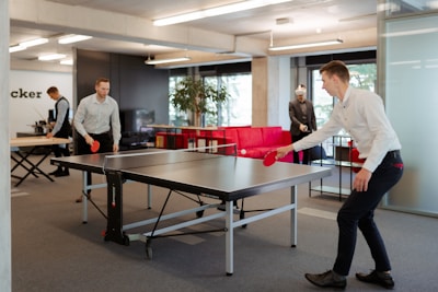 Two individuals are engaged in a game of table tennis in an office setting. The room is well-lit with modern furnishings including a red couch and potted plants. In the background, there are other people working at desks and someone wearing a VR headset.
