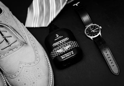 Elegantly arranged on a dark surface, a pair of stylish leather brogue shoes sits alongside a sophisticated wristwatch. Next to these items is a bottle of cologne with a checkered design and a striped necktie, suggesting a theme of classic men's fashion.