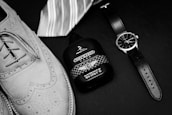 Elegantly arranged on a dark surface, a pair of stylish leather brogue shoes sits alongside a sophisticated wristwatch. Next to these items is a bottle of cologne with a checkered design and a striped necktie, suggesting a theme of classic men's fashion.