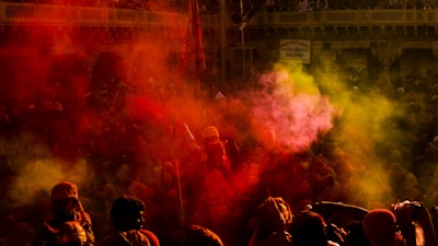 A vibrant festival scene filled with people celebrating, surrounded by clouds of colorful powder in red and yellow hues. The crowd is dense and lively, capturing the essence of a traditional cultural event, with a backdrop of ornately decorated buildings.