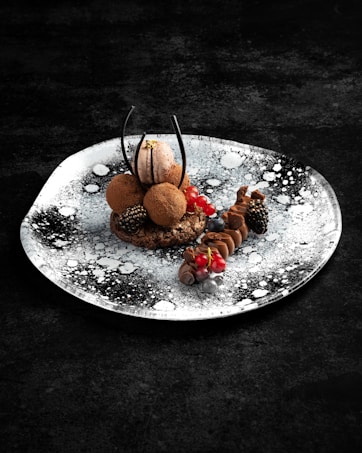An artistic dessert presentation on a plate with a black and white splatter design. The dessert consists of a chocolate macaron, truffles, and a variety of berries including blackberries and red currants, arranged with chocolate decorations.