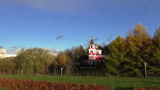 A vibrant red and white church with a prominent spire stands amidst a backdrop of lush green trees and a clear blue sky. The building is ornate, with crosses on top, and is set in a serene park environment with well-maintained lawns and shrubbery. Some residential buildings are visible in the distance, adding an urban touch to the natural setting.