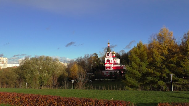 A vibrant red and white church with a prominent spire stands amidst a backdrop of lush green trees and a clear blue sky. The building is ornate, with crosses on top, and is set in a serene park environment with well-maintained lawns and shrubbery. Some residential buildings are visible in the distance, adding an urban touch to the natural setting.