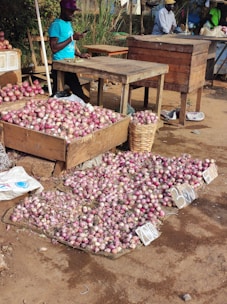 A street market with wooden tables and crates filled with red onions. A vendor in a blue shirt is standing beside the onions, using a phone. The onions are spread out on the ground with price tags visible. The setting appears outdoors with some greenery in the background.