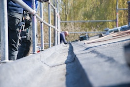 A person is working on a roof, surrounded by scaffolding. Several tools are visible, including a hammer hanging from a tool belt. The focus is on the textured surface of the roof.