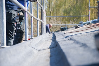 A person is working on a roof, surrounded by scaffolding. Several tools are visible, including a hammer hanging from a tool belt. The focus is on the textured surface of the roof.