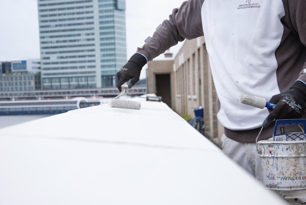 A person wearing a two-tone jacket and black gloves is painting a surface with a white roller brush. There is a can of paint resting on the structure nearby, which has paint splatters on it. The background shows a modern cityscape with tall buildings and overcast skies.