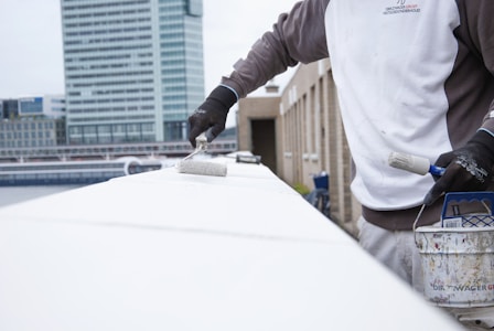 A person wearing a two-tone jacket and black gloves is painting a surface with a white roller brush. There is a can of paint resting on the structure nearby, which has paint splatters on it. The background shows a modern cityscape with tall buildings and overcast skies.