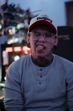 A person with glasses and stretched earlobes sticks out their tongue. They are wearing a cap with 'DadGang' inscribed on it. The background appears blurred with colorful lights, suggesting an indoor setting.