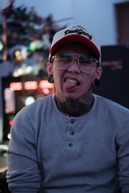 A person with glasses and stretched earlobes sticks out their tongue. They are wearing a cap with 'DadGang' inscribed on it. The background appears blurred with colorful lights, suggesting an indoor setting.