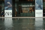 A modern storefront with large glass windows and two towering advertisements on either side. The left advertisement features the name 'ZARA' in bold black letters along with an image of a handbag and shoes. The right advertisement has text that reads 'NOW OPEN' and features a part of an outfit including a skirt and boots. In front of the store, there is a man walking while talking on a phone. The foreground displays a shallow body of water with reflections.