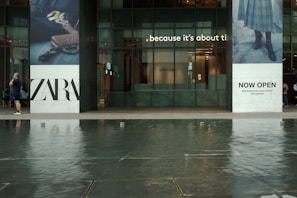 A modern storefront with large glass windows and two towering advertisements on either side. The left advertisement features the name 'ZARA' in bold black letters along with an image of a handbag and shoes. The right advertisement has text that reads 'NOW OPEN' and features a part of an outfit including a skirt and boots. In front of the store, there is a man walking while talking on a phone. The foreground displays a shallow body of water with reflections.