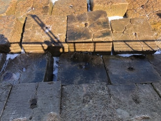 Several large concrete blocks arranged in a row, with visible cracks and erosion on their surfaces. The blocks are illuminated by sunlight, casting a distinct shadow of a metal railing across them. Some patches of white substance, likely salt or foam, are scattered on and between the blocks.