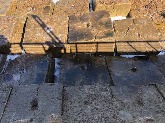 Several large concrete blocks arranged in a row, with visible cracks and erosion on their surfaces. The blocks are illuminated by sunlight, casting a distinct shadow of a metal railing across them. Some patches of white substance, likely salt or foam, are scattered on and between the blocks.
