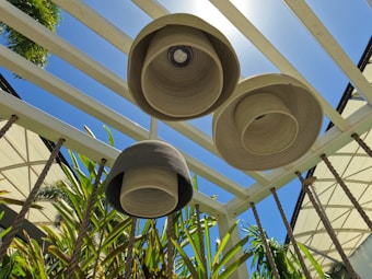 Three woven lampshades hang from a pergola structure made of white beams, with a bright blue sky in the background. Lush green plants rise up to fill the view, creating a natural contrast against the geometric lines above.