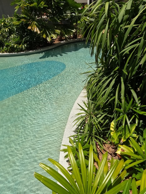 A serene, moody tropical poolside scene at dusk with soft green foliage, sky-blue water reflections, and natural wood decking inspired by Tulum and Greek coastal vibes.