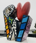 A colorful abstract sculpture composed of geometric shapes with various patterns, including a large red heart shape at the top and a structure with a peace symbol cutout. The piece features bold black outlines and a mix of polka dots, stripes, and solid colors.