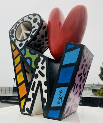 A colorful abstract sculpture composed of geometric shapes with various patterns, including a large red heart shape at the top and a structure with a peace symbol cutout. The piece features bold black outlines and a mix of polka dots, stripes, and solid colors.
