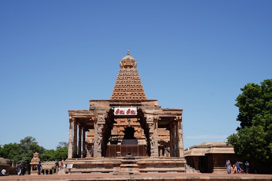 A historic temple with intricate carvings on its facade and a prominent pyramid-shaped tower. The temple is made of stone and is set against a clear blue sky. There are people walking around, dressed in colorful traditional attire, and the surroundings are lush with green trees.