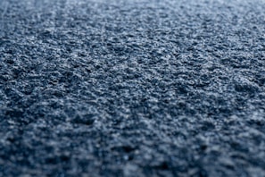 A close-up view of a rough, textured surface that appears to be asphalt. The surface is detailed with small stones and granules and is illuminated by soft light, creating a sense of depth and texture.