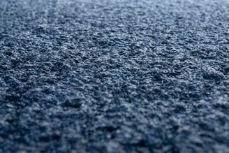 A close-up view of a rough, textured surface that appears to be asphalt. The surface is detailed with small stones and granules and is illuminated by soft light, creating a sense of depth and texture.