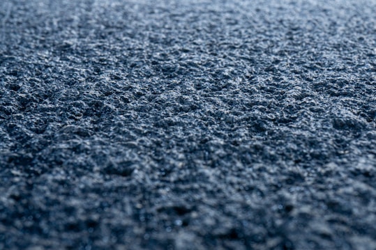 A close-up view of a rough, textured surface that appears to be asphalt. The surface is detailed with small stones and granules and is illuminated by soft light, creating a sense of depth and texture.