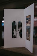 A handmade card is open to reveal intricate cut-out silhouettes of two figures standing in an arched window shape. On the opposite side, there's an artistic print depicting a scenic view with a dome and clouds, possibly inspired by a historical or famous building. The card is resting on a wooden surface, and the background is softly blurred.