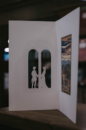 A handmade card is open to reveal intricate cut-out silhouettes of two figures standing in an arched window shape. On the opposite side, there's an artistic print depicting a scenic view with a dome and clouds, possibly inspired by a historical or famous building. The card is resting on a wooden surface, and the background is softly blurred.