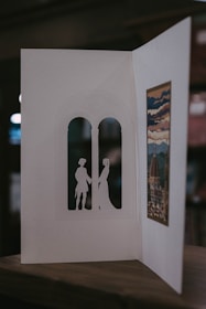 A handmade card is open to reveal intricate cut-out silhouettes of two figures standing in an arched window shape. On the opposite side, there's an artistic print depicting a scenic view with a dome and clouds, possibly inspired by a historical or famous building. The card is resting on a wooden surface, and the background is softly blurred.