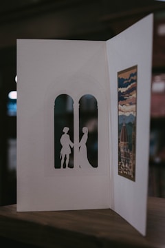 A handmade card is open to reveal intricate cut-out silhouettes of two figures standing in an arched window shape. On the opposite side, there's an artistic print depicting a scenic view with a dome and clouds, possibly inspired by a historical or famous building. The card is resting on a wooden surface, and the background is softly blurred.
