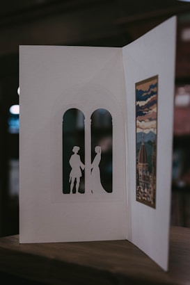 A handmade card is open to reveal intricate cut-out silhouettes of two figures standing in an arched window shape. On the opposite side, there's an artistic print depicting a scenic view with a dome and clouds, possibly inspired by a historical or famous building. The card is resting on a wooden surface, and the background is softly blurred.