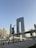 A modern urban landscape featuring a distinctive U-shaped skyscraper with a unique architectural design and multiple smaller buildings surrounding it. A bridge with people walking crosses over a waterway, and the scene is set against a clear blue sky.