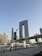A modern urban landscape featuring a distinctive U-shaped skyscraper with a unique architectural design and multiple smaller buildings surrounding it. A bridge with people walking crosses over a waterway, and the scene is set against a clear blue sky.
