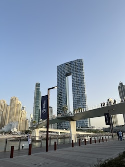A modern urban landscape featuring a distinctive U-shaped skyscraper with a unique architectural design and multiple smaller buildings surrounding it. A bridge with people walking crosses over a waterway, and the scene is set against a clear blue sky.