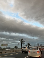 A busy road with multiple cars waiting in traffic, surrounded by palm trees. The sky is overcast with heavy, gray clouds, revealing a patch of blue sky. A street sign indicates 'Al Khail St' and there are buildings visible in the background.