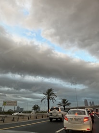 A busy road with multiple cars waiting in traffic, surrounded by palm trees. The sky is overcast with heavy, gray clouds, revealing a patch of blue sky. A street sign indicates 'Al Khail St' and there are buildings visible in the background.