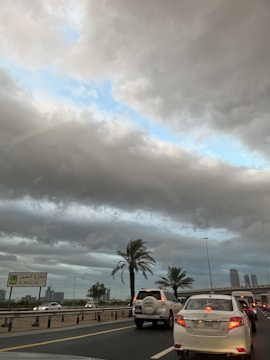 A busy road with multiple cars waiting in traffic, surrounded by palm trees. The sky is overcast with heavy, gray clouds, revealing a patch of blue sky. A street sign indicates 'Al Khail St' and there are buildings visible in the background.