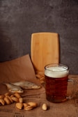 A glass of dark beer with foam sits on top of a newspaper, accompanied by almonds and dried fish. A wooden cutting board is positioned in the background against a gray textured wall.