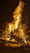 A large, intense fire with flames rising high into the air, surrounded by burning logs and branches. The scene is set against a dark background, highlighting the bright yellows and oranges of the flames.