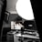A photography or video recording studio setup with a large softbox light providing illumination. A desktop setup with dual monitors is noticeable, one screen displaying a monochrome image of a woman. A professional microphone is placed in front of the monitors, suggesting a content creation or streaming environment. Various tech gadgets are arranged on and around the desk, including a control panel with multiple buttons and sliders, a keyboard, and a mouse.