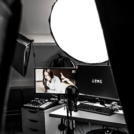 A photography or video recording studio setup with a large softbox light providing illumination. A desktop setup with dual monitors is noticeable, one screen displaying a monochrome image of a woman. A professional microphone is placed in front of the monitors, suggesting a content creation or streaming environment. Various tech gadgets are arranged on and around the desk, including a control panel with multiple buttons and sliders, a keyboard, and a mouse.