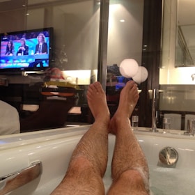 A pair of legs are seen stretched out in a bathtub filled with water. The room is dimly lit, and a television mounted on the wall is playing a show with three people sitting at a panel. The setting appears to be cozy, with reflections visible on a glass partition, hinting at a modern bathroom design.