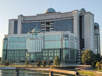 A large, modern building with a mix of neo-classical and modern architectural styles, featuring multiple domes and large glass windows. The structure is surrounded by trees and situated near a body of water, with a railing visible in the foreground.