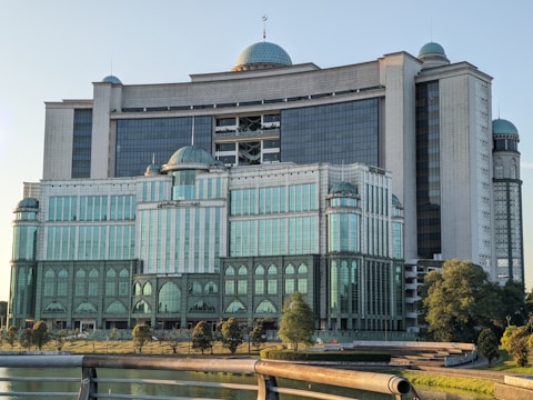 A large, modern building with a mix of neo-classical and modern architectural styles, featuring multiple domes and large glass windows. The structure is surrounded by trees and situated near a body of water, with a railing visible in the foreground.