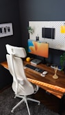A modern home office setup featuring a wooden desk with a computer monitor displaying colorful graphics. A sleek white office chair stands in front, and the wall is adorned with a pegboard holding various items, including a notebook and decorative elements. On the desk are a keyboard, mouse, potpourri sticks, a small plant, and a computing device.