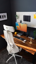 A modern home office setup featuring a wooden desk with a computer monitor displaying colorful graphics. A sleek white office chair stands in front, and the wall is adorned with a pegboard holding various items, including a notebook and decorative elements. On the desk are a keyboard, mouse, potpourri sticks, a small plant, and a computing device.