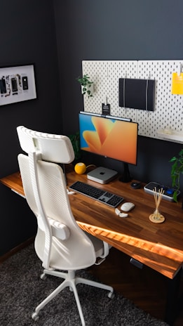 A modern home office setup featuring a wooden desk with a computer monitor displaying colorful graphics. A sleek white office chair stands in front, and the wall is adorned with a pegboard holding various items, including a notebook and decorative elements. On the desk are a keyboard, mouse, potpourri sticks, a small plant, and a computing device.
